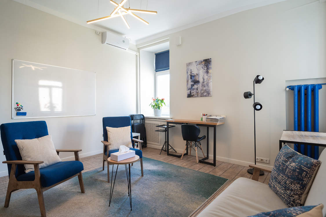 Два синих кресла и светлый диван вокруг столика, у окна рабочий стол и настенная доска.