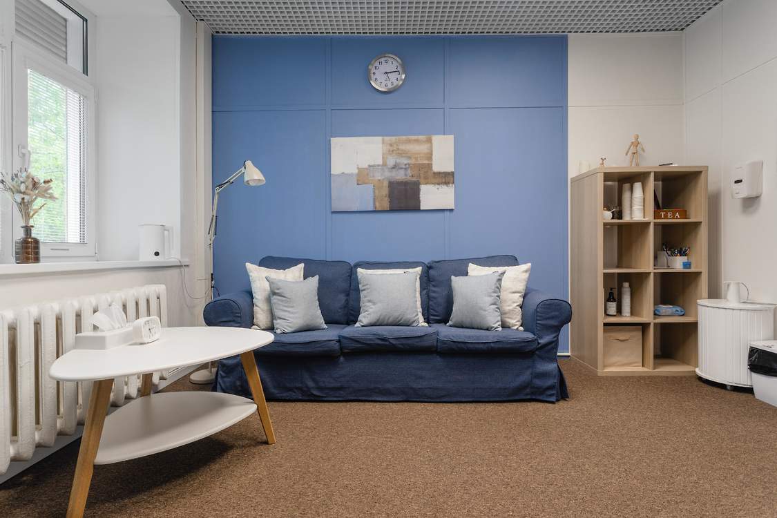 Синий диван под картиной у голубой стены, рядом белый столик и деревянный стеллаж.